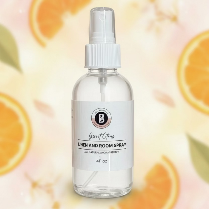 A clear glass spray bottle filled with white liquid, featuring a white pump top and a white label that reads 'Sweet Citrus LINEN AND ROOM SPRAY.' The bottle is placed on a white marble surface with a softly blurred indoor background showing plants and a doorway.