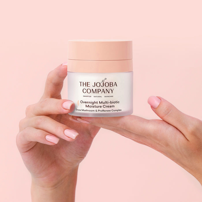 A person's hands holding a jar of The Jojoba Company Overnight Multi-biotic Moisture Cream against a soft pink background.