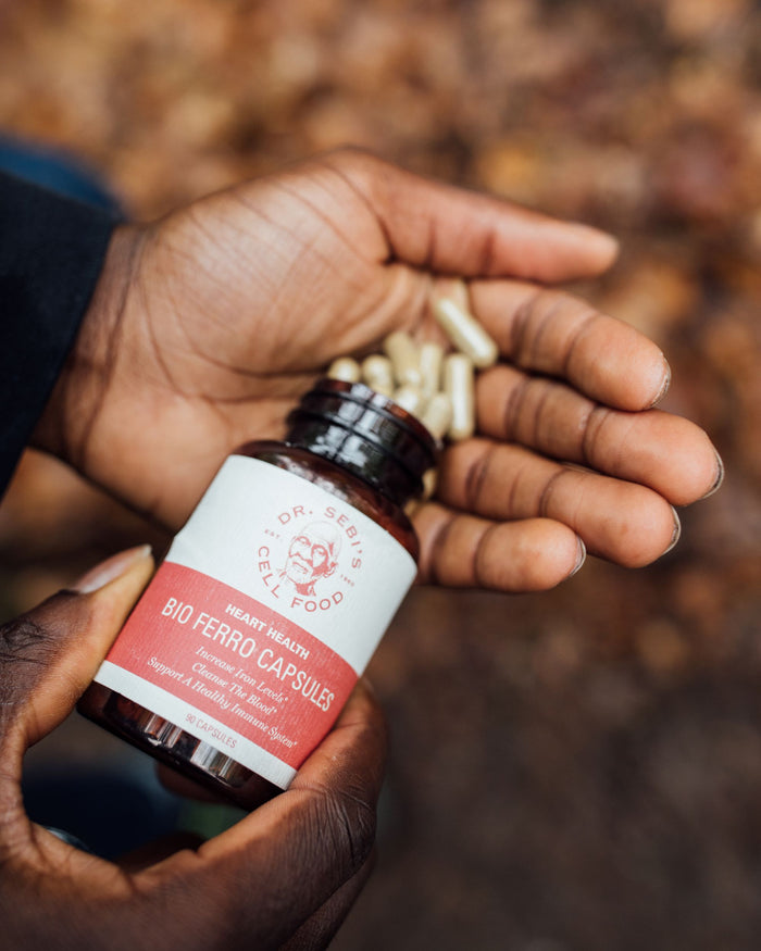 Hand holding Dr. Sebi's Bio Ferro Capsules for heart health with loose capsules above against natural blurred background.