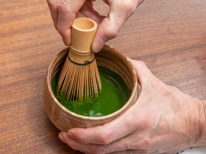 Person using a bamboo whisk to stir green tea in a ceramic bowl on a wooden surface. Koicha