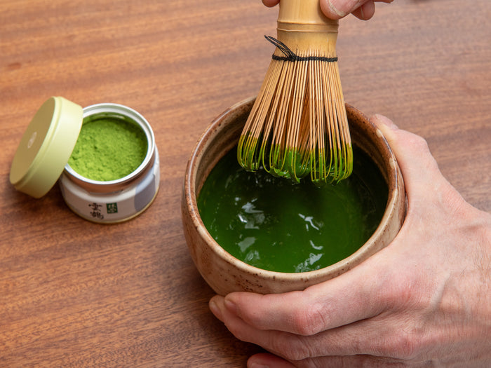 Person whisking green tea in a bowl with a container of matcha powder on a wooden surface. Koicha