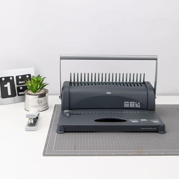 Black office binding machine on a gray cutting mat with a small plant and calendar in the background.