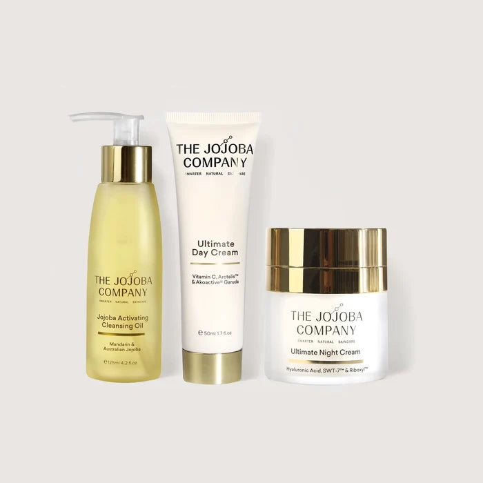 Three skincare products from The Jojoba Company arranged on a light gray background: a yellow cleansing oil bottle with a gold pump, a white day cream tube with a gold base, and a white night cream jar with a gold lid.