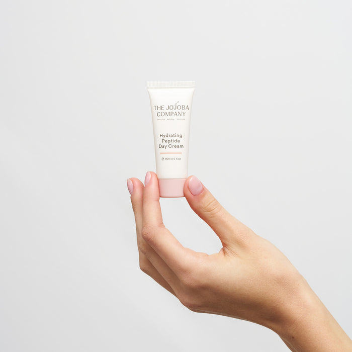 Hand holding a tube of The Jojoba Company hydrating peptide day cream against a light gray background