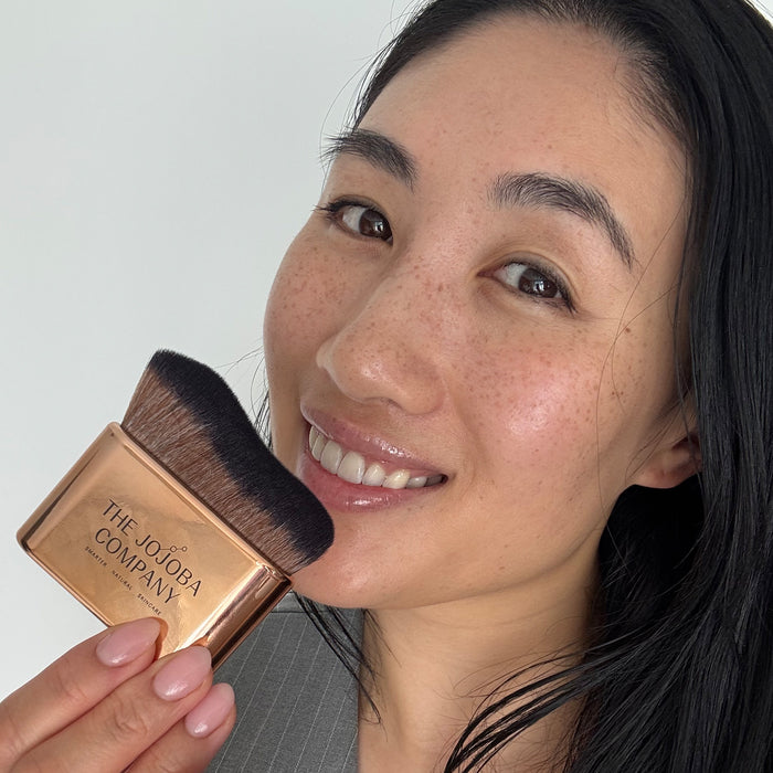 A close-up lifestyle shot of a woman holding a makeup brush from The Jujuba Company next to her cheek, showing the rose gold and black brush against a clean white background.