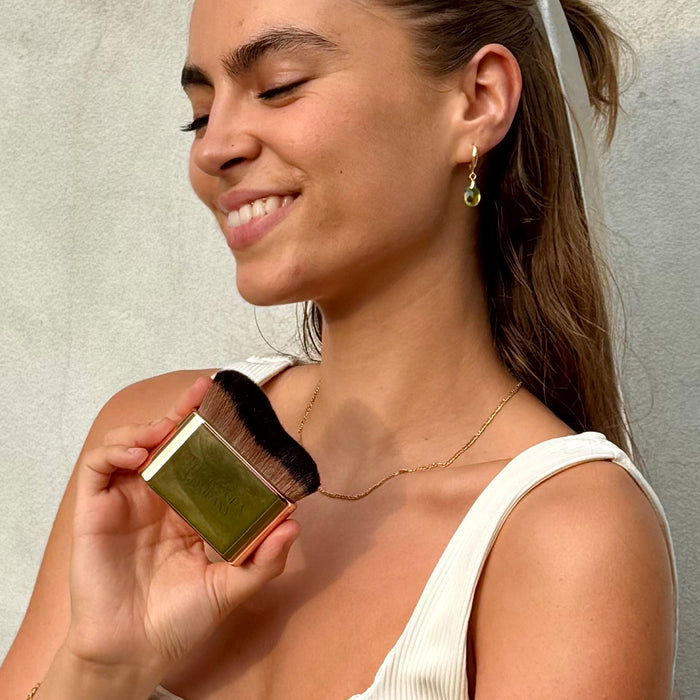 A woman in a white ribbed tank top holds a small square makeup compact with a gold-toned case and dark bristles, wearing a gold necklace and earring against a light gray textured wall.