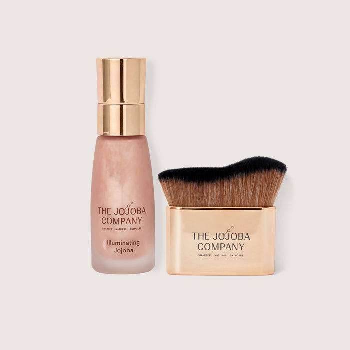 The Jojoba Company Illuminating Jojoba cosmetic product shown as a frosted bottle with a metallic gold cap next to a matching square makeup brush with a gold-toned base and black-and-brown bristles, on a light gray background.
