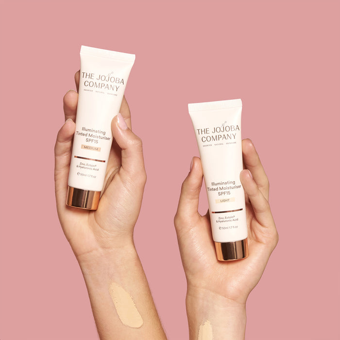 Two hands holding The Jojoba Company skincare products against a pink background