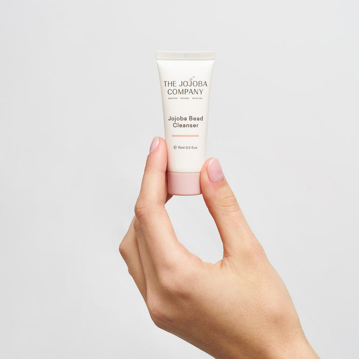 Hand holding a tube of Jojoba Bead Cleanser from The Jojoba Company against a light gray background