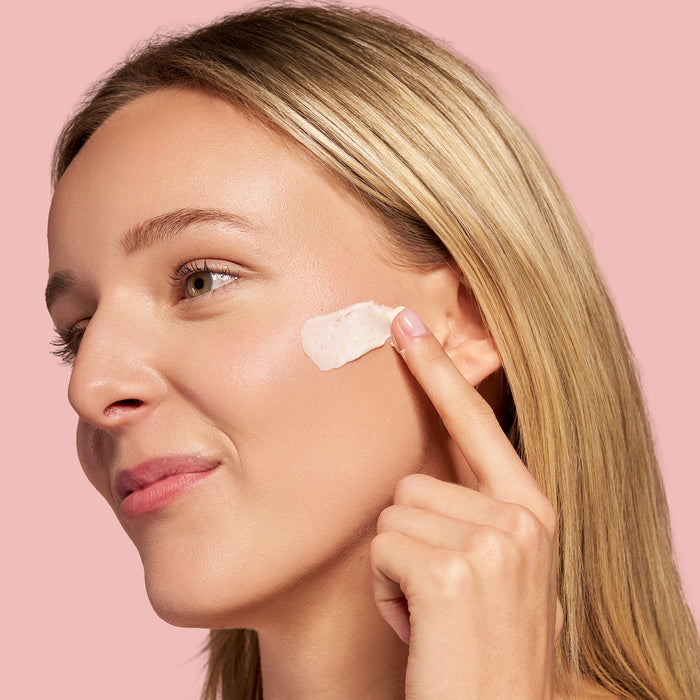 Model applying Jojoba Bead Cleanser from The Jojoba COmpany to her face with a pink background