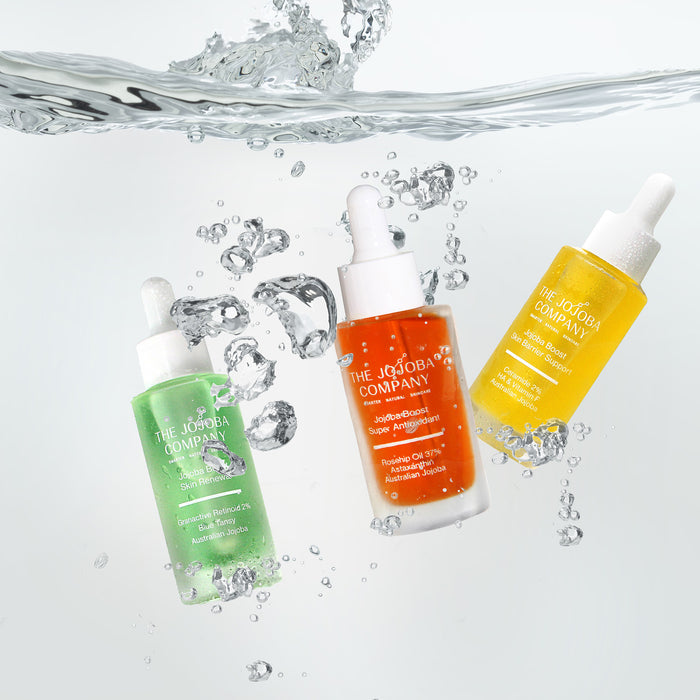 Three serum skincare bottles from The Jojoba Company with water droplets and splashes on a light gray background.