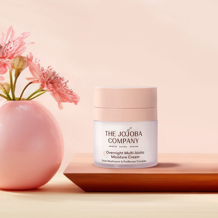 A jar of The Jojoba Company Overnight Multi-biotic Moisture Cream sits on a wooden tray next to a pink vase with flowers, set against a soft peach-pink background.