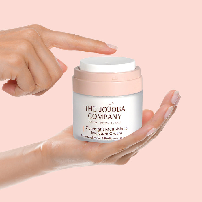 A hand holding a jar of The Jojoba Company Overnight Multi-biotic Moisture Cream against a solid pale pink background.